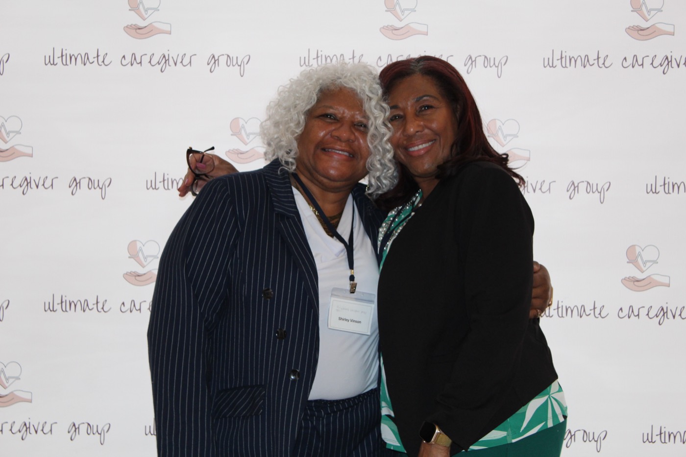 Two attendees embrace at the Ultimate Caregiver Group backdrop.